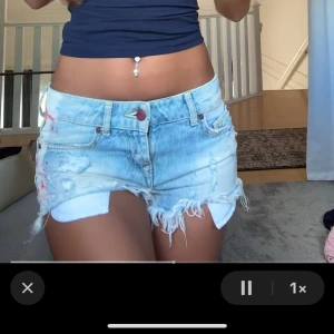 Lågmidjade ljusblå jeansshorts med fransar - Säljer ett par riktigt snygga lågmidjade jeansshorts i ljusblå denim. De har slitna detaljer, fransig kant och klassisk femficksmodell. Shortsen har en röd knapp och dragkedja framtill. Perfekta för dig som gillar en chill och trendig look på sommaren.