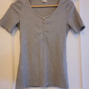 Grå ribbad topp med dragkedja XS - Grå ribbad topp från Cubus i storlek XS men passar S om man gillar lite mer kroppsnära passform. Modellen har korta ärmar, rund halsringning och en cool silverfärgad dragkedja framtill med ringdetalj.