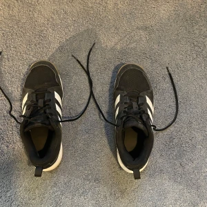 Svarta Adidas sneakers med vita ränder - Snygga svarta Adidas sneakers med klassiska vita ränder på sidorna och gummisula i brunt. Ovandelen är i textil och syntet, med snörning framtill och rund tå. Perfekta inneskor.