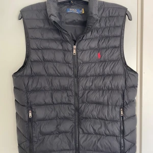 Ralph Lauren väst - Snygg svart dunväst från Polo Ralph Lauren i nyskick. Västen har två fickor med dragkedja och klassisk röd broderad logga på bröstet. Aldrig använd och den är i regular fit! Den är även justerbar i midjan runtom med resor så går att fixa så den passar en själv. Skick 10/10 helt ny och bara att höra av sig vid frågor eller funderingar!