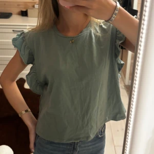 Grön blus med volangärmar - Snygg grön blus med volangdetaljer på ärmarna. Modellen är ärmlös och har en avslappnad passform, perfekt till jeans eller kjol. Tillverkad i ett mjukt bomullstyg som känns skönt mot huden. Enkel och stilren design som lyfter din outfit.
