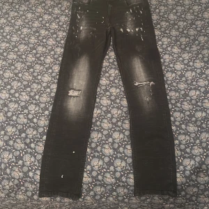 Svarta slitna jeans med detaljer - Säljer ett par svarta jeans med coola slitningar och färgstänk på benen. Jeansen har raka ben och en något tvättad look med trasiga detaljer på båda benen. Perfekta för dig som gillar en edgy streetstil.