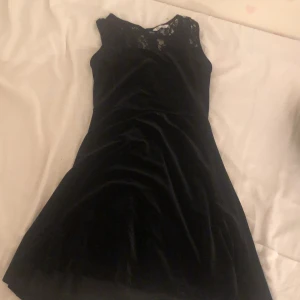 Svart sammetsklänning med spets - Supersnygg svart sammetsklänning med spetsdetaljer upptill. Ärmlös modell med rund halsringning och lätt utställd kjol. Perfekt för dig som vill ha en elegant och stilren look med en touch av lyxig känsla.