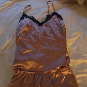 Rosa satin linnetopp samt shorts med spets pyjamas - Säljer en glansig rosa linnetopp samt shorts pyjamas i satin med tunna axelband och svart spetsdetalj. Perfekt för dig som gillar en chic och feminin stil. Pyjamasen har en mjuk, följsam känsla och passar snyggt till dig som vill vara lite extra fin på natten också.