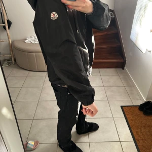 Svart vindjacka från Moncler - Snygg svart vindjacka från Moncler med huva och dragkedja. Jackan har Moncler-logga på ärmen och detaljer i rött och vitt på huvan. Perfekt för blåsiga dagar. Den är använd några gånger och säljs då den inte passar Hål i ficka och armhåla! Pris kan diskuteras!