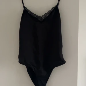 Svart body med spets från Zara - Supersnygg svart body från Zara med tunna axelband och v-ringning dekorerad med spets. Sidenliknande material som ger en lyxig känsla. Perfekt att styla med jeans eller kjol för en trendig look. Aldrig använd på grund av att den är för liten på mig.