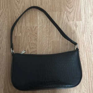 Väska - svart axelväska från H&M Divided med croco-mönstrad yta. Väskan har en enkel silvrig dragkedja och  svart axelrem. 