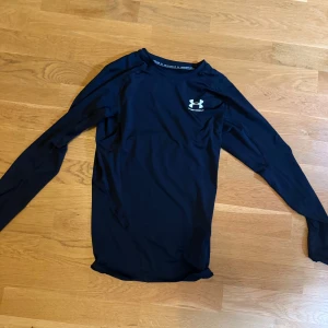 Svart långärmad träningströja Under Armour compression  - Svart långärmad träningströja från Under Armour med logga på bröstet. Tight passform och tillverkad i ett lätt, syntetiskt material som andas. Perfekt för gymmet eller löprundan. Enkel och stilren design som funkar till all sport.
