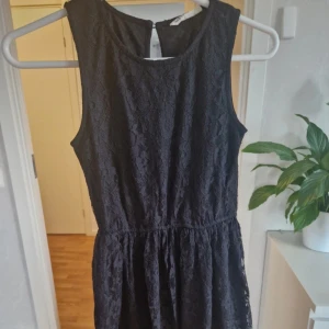 Svart spetsklänning från KappAhl - Supersnygg svart kortklänning från KappAhl med blommig spets och ärmlös design. Klänningen har rund halsringning och en liten knapp i nacken. Perfekt för dig som gillar klassisk stil med en twist.