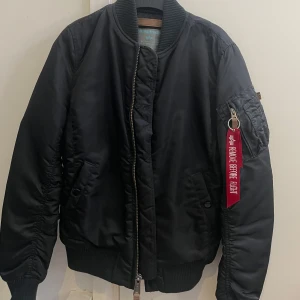 Svart bomberjacka Alpha Industries XS - Säljer en klassisk svart bomberjacka från Alpha Industries i storlek XS. Jackan har orange foder, dragkedja framtill och ikonisk röd tag på ärmen. Ribbstickade muddar och flera fickor ger en cool och tidlös look. Perfekt för dig som gillar streetstyle. Pris kan diskuteras. 