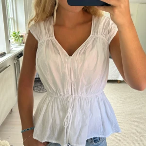 Vit blus med knyt och v-ringning - Söt vit blus med v-ringning och knyt framtill. Modellen har korta ärmar och markerad midja med resår och volangig nederdel. Perfekt till jeans för en fräsch och somrig look.