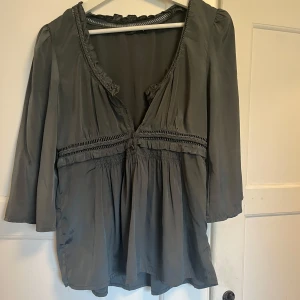 Grå blus med volang och dragkedja - Supersnygg grå blus i glansigt material med v-ringning, volangkant och dekorativa knappar framtill. Blusen har trekvartsärm, smockdetaljer under bysten och dragkedja i sidan för extra passform. Perfekt till jeans eller kjol för en chill look.