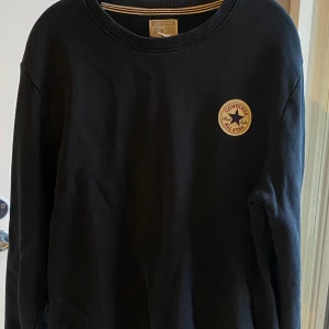 Tröja från Converse - Tröja från Converse med klassisk rund hals och lång ärm. På bröstet sitter en broderad Converse All Star-patch i vitt, rött och blått. Tröjan är i mjuk bomull och har en avslappnad passform, perfekt för en chill och sportig stil. 