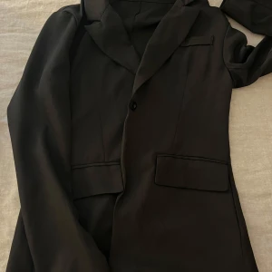 Svart klassisk kavaj med fickor - Stilren svart kavaj med klassisk krage och två framfickor med lock. Enkelknäppt framtill och diskret bröstficka. Perfekt för en clean och tidlös look. Passar dig som gillar minimalistisk stil.