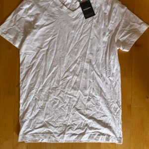 Vit v-ringad t-shirt från Dressmann - Klassisk vit t-shirt från Dressmann med v-ringning. Tillverkad i mjuk bomull och Fairtrade-märkt. Enkel och stilren design utan tryck eller mönster, perfekt för en clean look. Kortärmad modell som passar året runt.