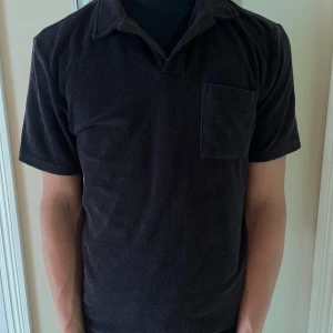 Svart frotté pikétröja East West - Svart pikétröja från East West i mjuk frotté. Klassisk krage, kort ärm och bröstficka ger en chill vibe. Perfekt för dig som gillar en enkel men ändå unik look. Passar till både jeans och shorts.