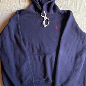 Marinblå hoodie från Polo Ralph Lauren - Säljer en marinblå hoodie från Polo Ralph Lauren med vit snörning och klassisk röd logga broderad på bröstet. Tröjan har huva, känguruficka och långa ärmar. Perfekt för dig som gillar stilren och avslappnad look.