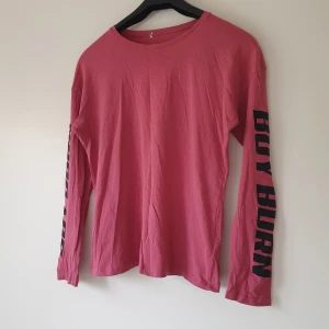 Rosa långärmad tröja med tryck från NA-KD strl M - Rosa långärmad tröja i bomull med svart texttryck 'BOY BURN' längs ena ärmen. Enkel och avslappnad passform med rund halsringning. Perfekt för dig som gillar streetstyle och vill ha något bekvämt men ändå snyggt.  Små noppror och litet hål vid nacken annars fint skick
