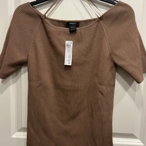 Ribbad mörk beige topp från Lindex - Snygg ribbad topp från Lindex i en varm brun nyans. Modellen har korta ärmar och en något bredare halsringning. Perfekt att styla med jeans eller kjol för en trendig look. Materialet är mjukt och stretchigt för en bekväm passform.