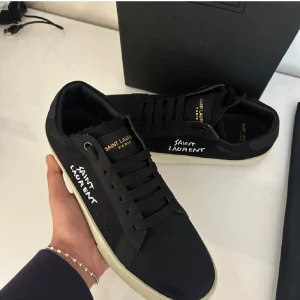 Svarta Saint Laurent sneakers - Svarta sneakers från Saint Laurent med vit logga på sidan och guldfärgad text på plösen. Skorna har svarta snören, vit platt sula och klassisk rund tå. Snygg minimalistisk design i tyg och skinn, levereras med originalkartong och dustbag.