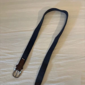 Mörkblå flätat bälte med skinnspänne - Snyggt mörkblått bälte med flätad design och detaljer i brunt skinn vid spännet. Spännet är i silverfärgad metall och bältet har en klassisk, stilren look som passar till jeans eller chinos.