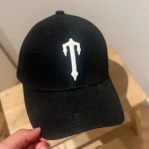 Svart keps från Trapstar - Svart keps från Trapstar med vit broderad logga framtill och texten 'TRAPSTAR' broderad baktill. Justerbar rem med metallspänne. Klassisk böjd skärm och stilren streetwear vibe.
