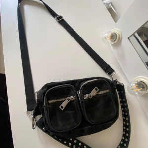 Svart mocka axelväska med nitar - Cool svart axelväska i mocka med två framfickor och silvriga dragkedjor. Väskan har en justerbar axelrem och en extra rem med silvriga nitar för en edgy vibe. Perfekt storlek för mobilen, plånboken och lite till. Enkel att matcha till streetwear eller rockiga outfits. Har några slitningar men är därför också väldigt prisvärd och fungerar precis lika bra.