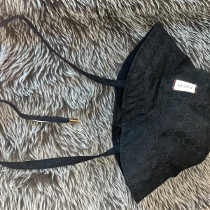 Svart bucket hat från Gucci - Snygg svart bucket hat från Gucci med diskret GG-monogram över hela hatten. Hatten har smala knytband med metalländar och en vit etikett med Gucci-logga framtill. Materialet känns slitstarkt och har en lyxig känsla, perfekt för att lyfta din outfit.