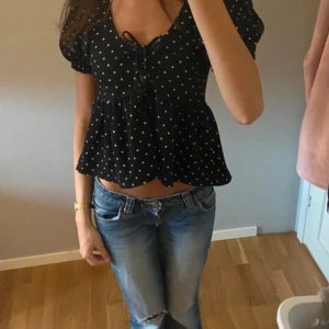 Svart prickig blus med knyt - Supersöt svart blus med vita prickar och korta puffärmar. Toppen har v-ringning med knyt framtill och volang nedtill som ger en luftig och trendig look. Perfekt till jeans för en chill och snygg outfit.