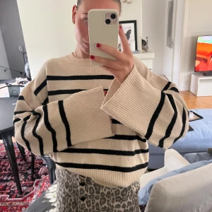 Randig beige stickad tröja - Säljer en oversized stickad tröja i beige med svarta ränder. Tröjan har breda ärmar och en avslappnad passform, perfekt för lager på lager. Mjuk och skön med klassisk rund halsringning. Superenkel att styla till både jeans och kjol.