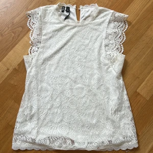 Vit spetsblus från Pieces - Superfin vit blus från Pieces med vackra spetsdetaljer och blommigt mönster. Blusen är ärmlös med dekorativa kanter och knapp i nacken. 