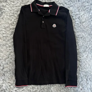 Svart långärmad pikétröja Moncler M - Svart långärmad pikétröja från Moncler med klassisk krage och knäppning framtill. Kragen och ärmsluten har snygga rödvita och marinblå ränder. Tröjan har Moncler-logga på bröstet och är gjord i bomull för en skön känsla.