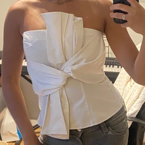 Vit bandeau topp med knytdetalj - Supersnygg vit bandeau korsett too H&M med en stor knytdetalj framtill. Toppen är axelbandslös och har dragkedja i ryggen för enkel påklädning. Köpt i vintras men aldrig kommit till användning, alltså helt ny.