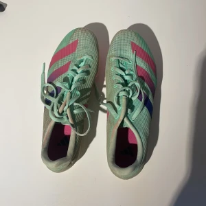 Mintgröna adidas spikskor strl 39⅓ - Snygga mintgröna adidas spikskor med rosa och blå ränder. Ovandel i syntetmaterial och snörning för bra passform. Sulan är rosa med metallspikar för maxat grepp på banan. Perfekta för friidrott och träning på bana.