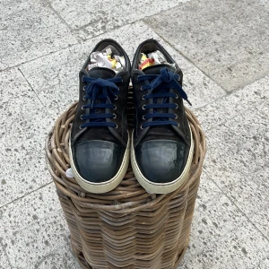 Svarta sneakers från Lanvin i mocka - Snygga svarta sneakers från Lanvin med ovandel i mocka och lackad tå. Skorna har mörkblå snörning och vit sula som ger en clean kontrast. Perfekta för dig som gillar stilrena och exklusiva sneakers med en twist.