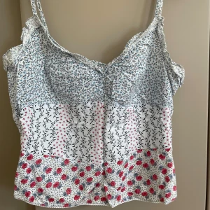 Blommig linnetopp med volangdetaljer - Supersöt linnetopp med smala axelband och volangdetaljer vid axlarna. Toppen har ett patchworkmönster med små blommor i blått, svart och rött på vit botten. Perfekt för sommarens alla tillfällen och ger en romantisk vibe. Dragkedja insidan för perfekt passform. 