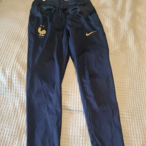 Blå Nike FFF träningsbyxor - Mörkblå träningsbyxor från Nike med guldiga detaljer. Byxorna har smal passform, resår och snörning i midjan samt muddar vid benslut. Fickor med dragkedja. Se bilder för mått. Se gärna även mina andra annonser och skapa bundles. 