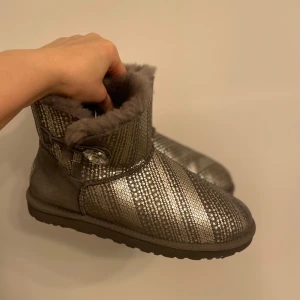 Glittriga boots från UGG - Snygga och varma boots från UGG i grå mocka med glittriga detaljer. De har en Swarovski dekorativ knapp på sidan och är fodrade med mjuk fårskinn päls för extra komfort. Perfekta för att hålla stilen under kalla dagar. Swarovski crystal bak också. Har använt en gång, som ny.
