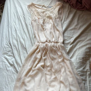 Champagnefärgad klänning från ASOS - Säljer en elegant champagnefärgad klänning från ASOS med spetsdetaljer vid halsen och en öppen rygg. Klänningen har en elastisk midja och är ärmlös, vilket ger en luftig och stilren look. Perfekt för sommarens festligheter!