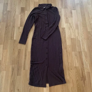 Brun ribbad långklänning från Happy Holly - Säljer en elegant brun långklänning från Happy Holly. Klänningen är ribbad med knappar längs hela framsidan och har långa ärmar. Perfekt för en stilren look.