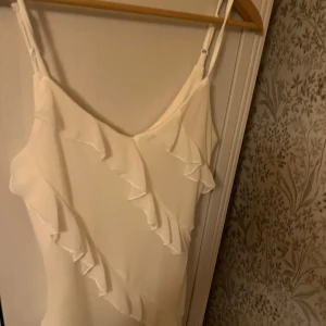 Vit klänning med volanger - Elegant vit klänning med volanger som ger en romantisk känsla. Klänningen har smala axelband och en figurnära passform. Perfekt för sommarens festligheter.