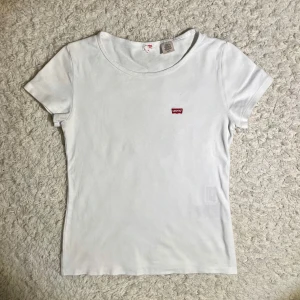 Vit t-shirt från Levi's - Säljer en klassisk vit t-shirt från Levi's med en liten röd logga på bröstet. T-shirten har en rund hals och korta ärmar, perfekt för en avslappnad stil. Passar till det mesta och är ett måste i garderoben!
