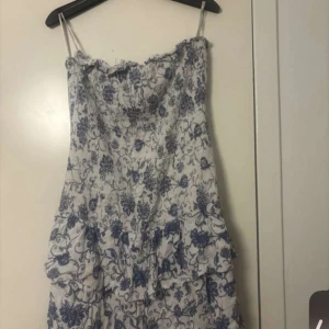 Blommig klänning med volanger - Charmig vit klänning med blått blommönster och volanger. Axelbandslös design som ger en somrig känsla. Perfekt för varma dagar.