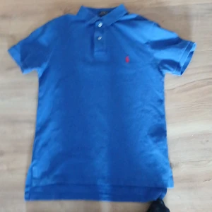 Blå pikétröja från Ralph Lauren - Säljer en klassisk blå pikétröja från Ralph Lauren med röd broderad logotyp på bröstet. Tröjan har korta ärmar och en stilren krage med knappar. Perfekt för en avslappnad och stilren look. Är ett liitet hål (på 5:e bilden) annars är den jättefin👍👍 Pris kan diskuteras👍👍