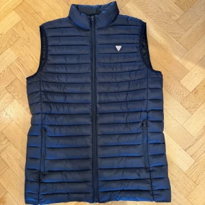Svart dunväst från Guess - Snygg svart dunväst från Guess med dragkedja och perfekt nu för våren. Västen har en stilren design med en liten detalj på bröstet. Använd fåtal gånger skulle säga skick 9/10, nypris ca 800 kr. Pris går att diskutera vid snabb affär💰 Vid frågor eller funderingar hör av er!