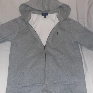 Grå hoodie från Polo Ralph Lauren - Säljer en klassisk grå hoodie från Polo Ralph Lauren med dragkedja och huva. Den har en liten broderad logga på bröstet och är tillverkad i mjukt material. Perfekt för en avslappnad stil.