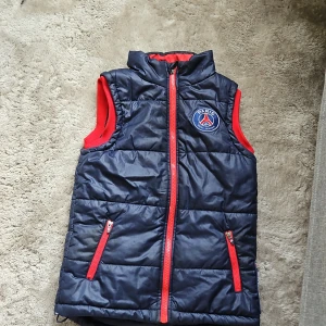 Blå dunväst från Paris Saint-Germain - Säljer en blå dunväst från Paris Saint-Germain med röda detaljer och dragkedja. Västen har klubbens logga på bröstet och är perfekt för fotbollsfans. Den är quiltad och har en sportig look. Har ett hack vid dragkedja men går och fixa med garn om man syr.