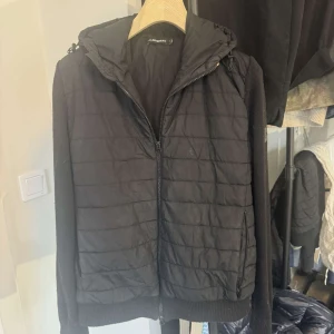 J.Lindeberg Cardigan - Snygg svart quiltad jacka från J.Lindeberg med huva och dragkedja. Jackan har långa ärmar och ribbade muddar. Enda deffekten är att det är hål i ena innerfickan. Perfekt för våren. Storlek M.