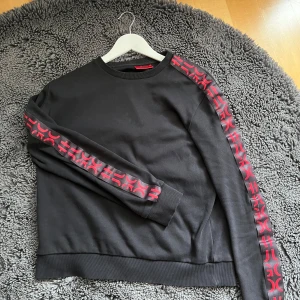 Svart tröja från Hugo Boss äkta - Snygg svart tröja från Hugo Boss med röda detaljer längs ärmarna. Tröjan har en rund halsringning och ribbade muddar vid ärmslut och nederkant. Perfekt för en stilren look. Passaer även xs. 