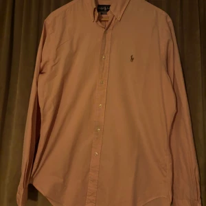 Rosa skjorta från Ralph Lauren - Snygg rosa skjorta från Ralph Lauren med klassisk krage och broderad logotyp på bröstet. Skjortan har långa ärmar och knappar framtill. Perfekt för en stilren look. 